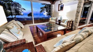 Sala de estar ampla com uma grande porta de vidro que dá para uma varanda com vista para o mar. Há um conjunto de sofás e uma mesa de centro no espaço.