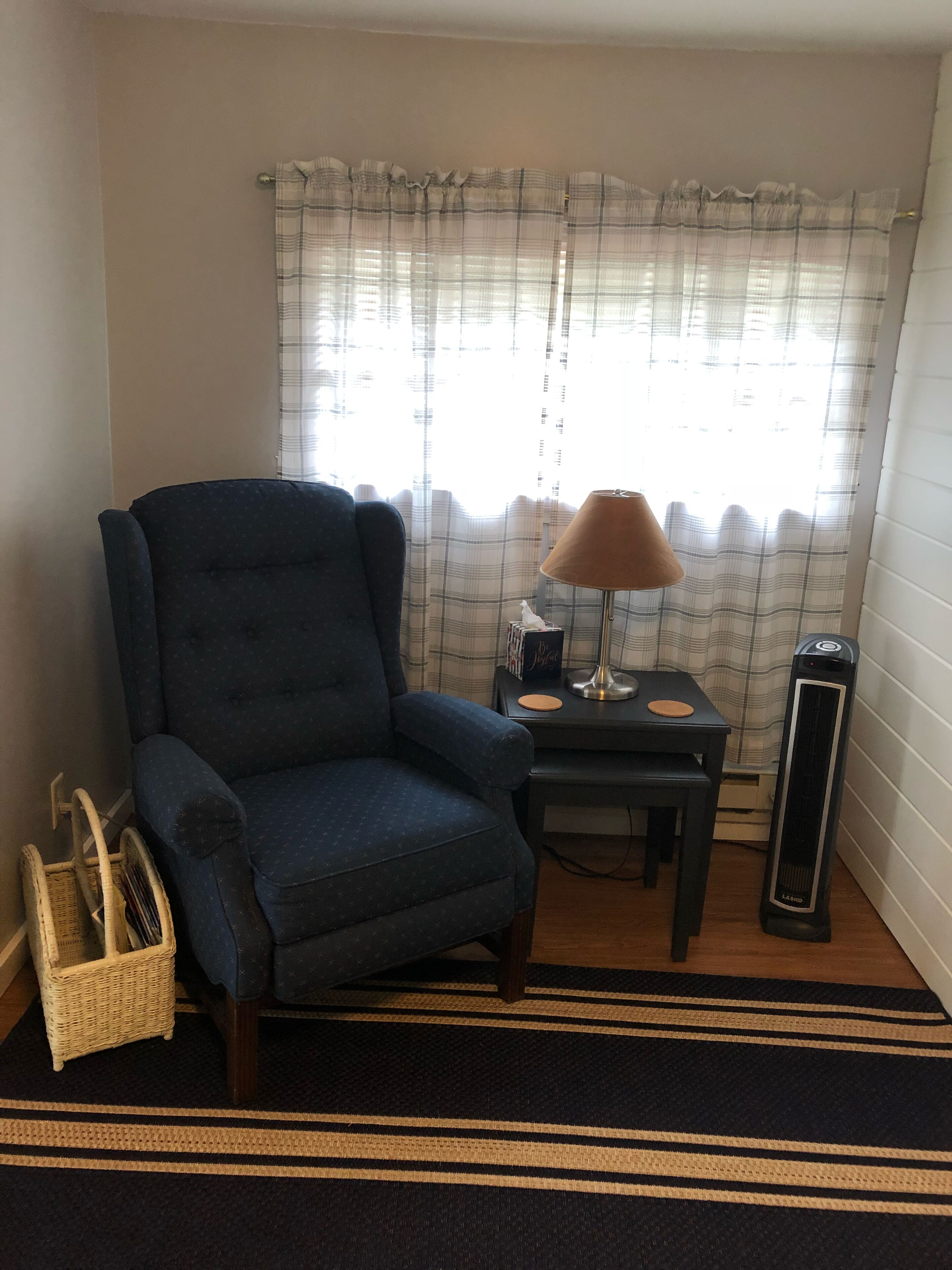 Une pièce avec un fauteuil bleu, une petite table avec une lampe, un radiateur et un panier en osier.