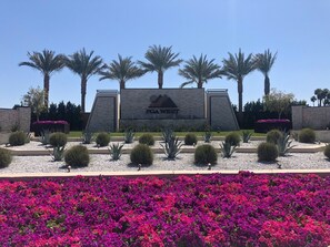Señal de PGA WEST con un jardín ajardinado al frente, que incluye palmeras y flores púrpuras.
