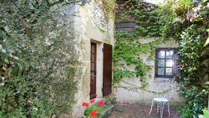 Un edificio rústico de piedra con muros cubiertos de hiedra, una puerta de madera, una ventana y una silla de metal blanco.