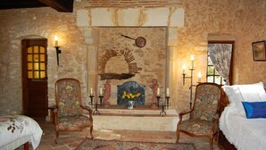 Una habitación rústica con paredes de piedra, una chimenea con un arco decorativo, dos sillas de madera, una cama con una colcha blanca y azul, y una alfombra estampada.
