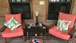 Deux fauteuils rouges, recouverts d’un tissu et agrémentés de coussins aux motifs tropicaux, une petite table avec une lampe, et une boîte étiquetée « ANTIQUITÉS » posée sur le sol.