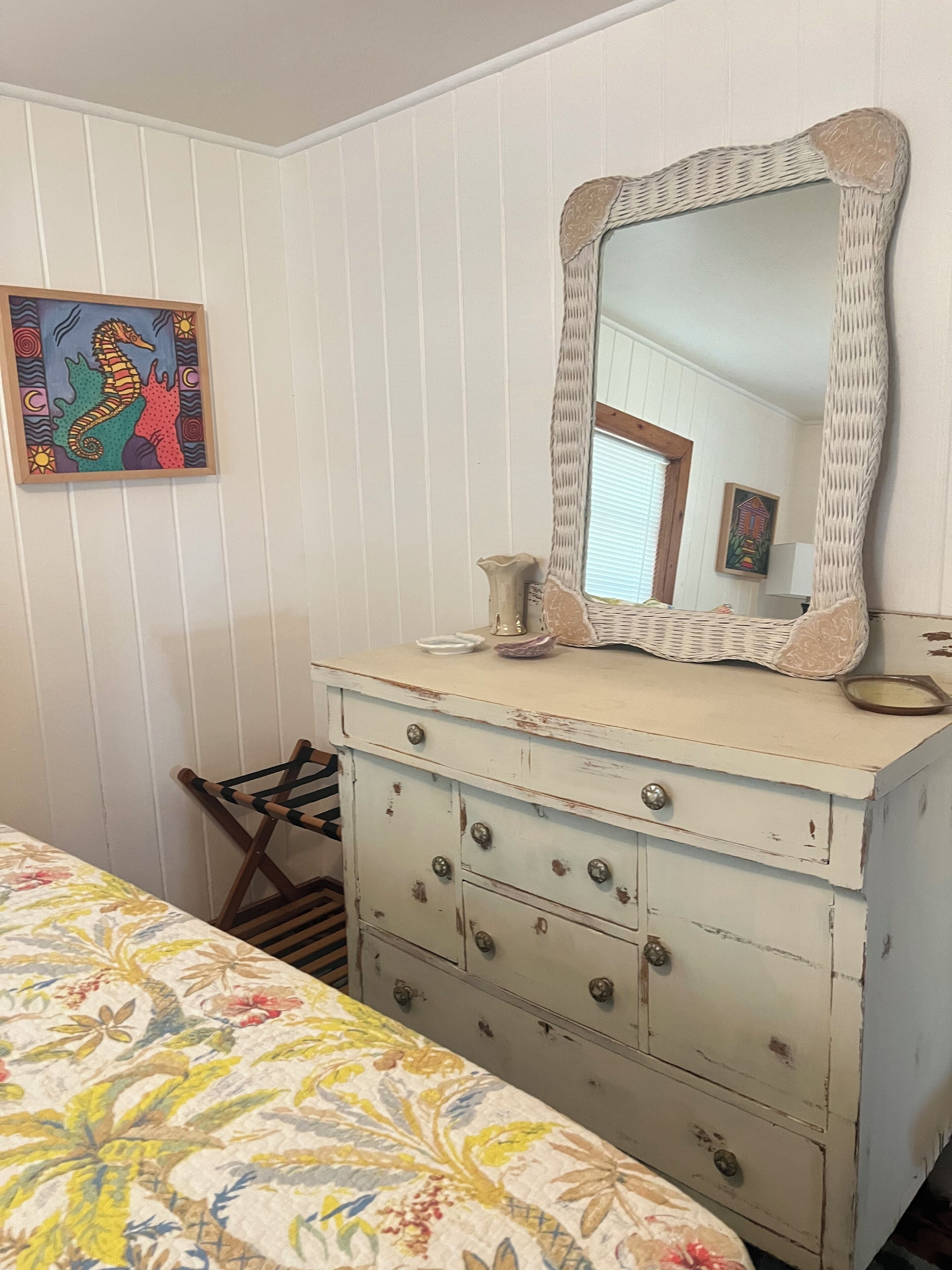 A room with a white dresser, a mirror, a floral bedspread, and a framed seahorse painting on the wall.