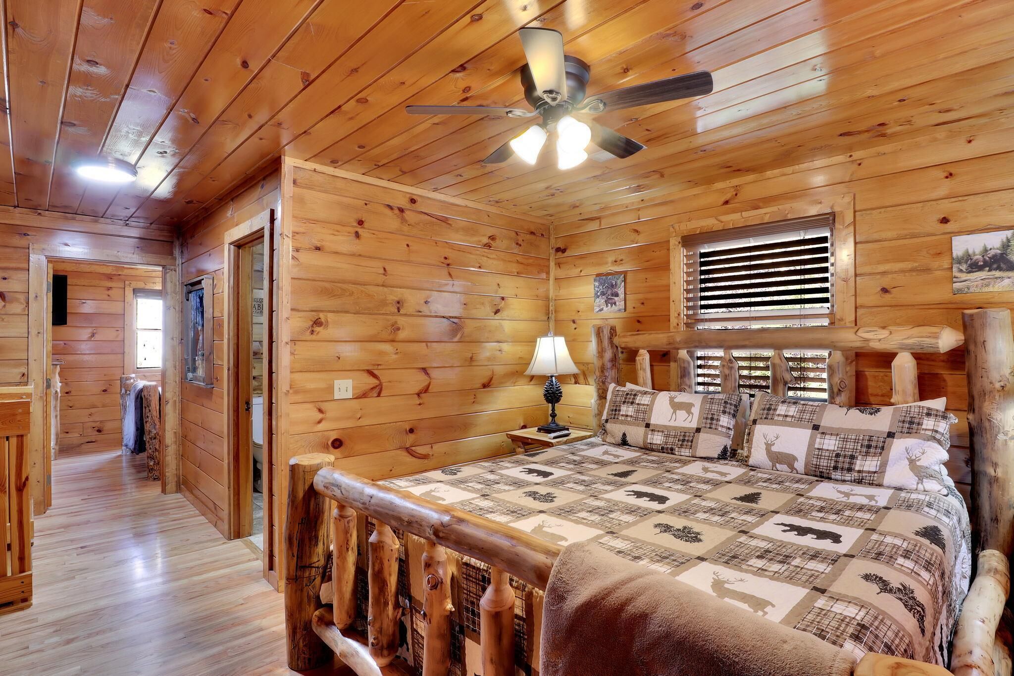 Une chambre dans un chalet en bois, avec un lit, un ventilateur de plafond, une fenêtre avec des stores et une rambarde en bois.
