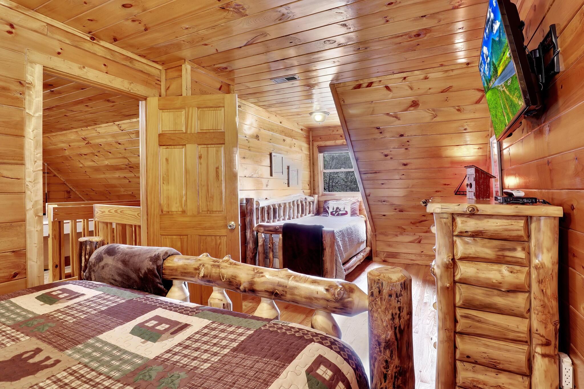 A cozy cabin room with a wooden ceiling, a bed with a quilt, a wooden chair, and a small table with a lamp.