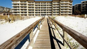 A wooden boardwalk leading to a multi-story hotel with balconies, situated on a sandy beach.