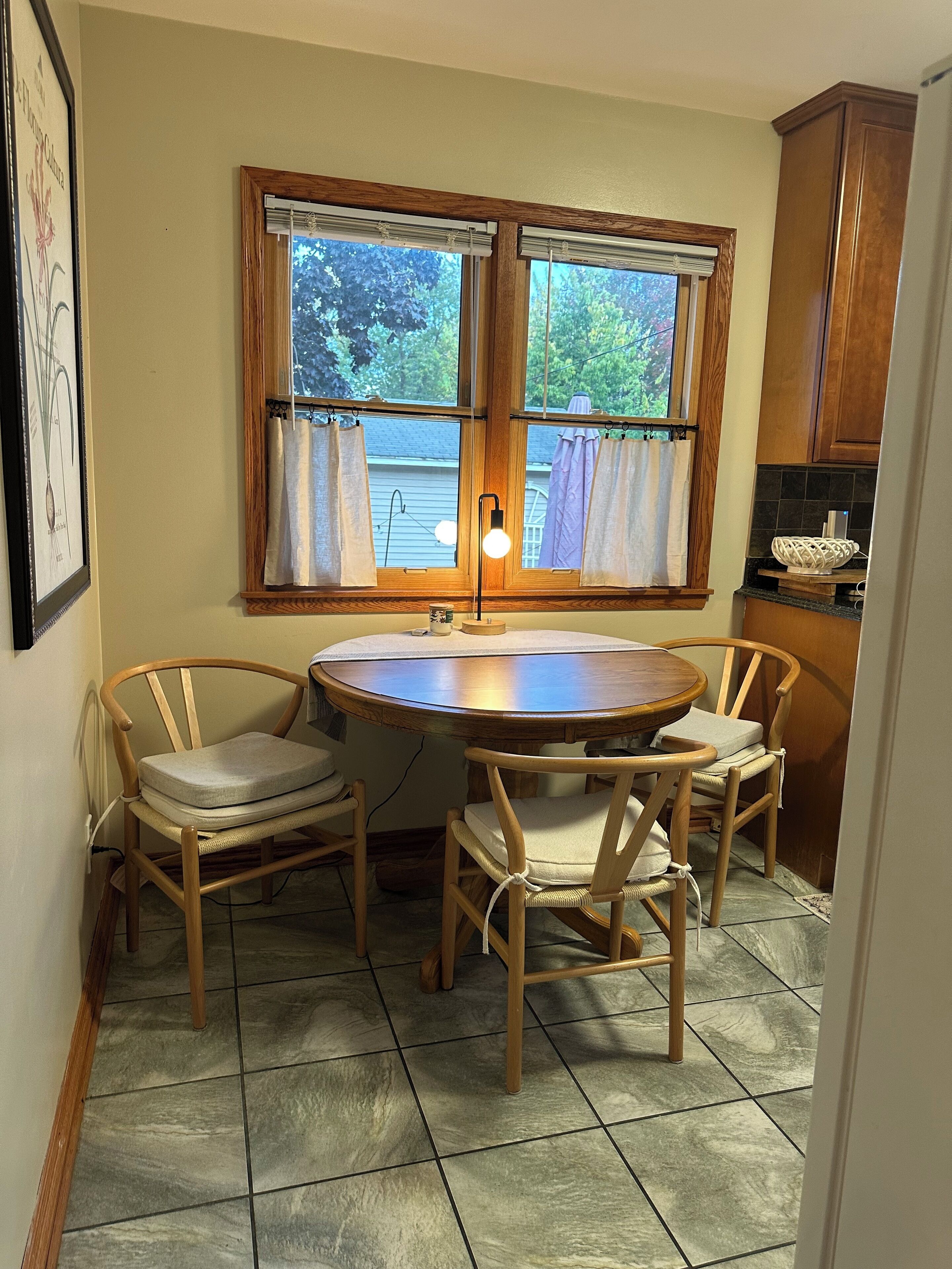A dining area with a round wooden table and four chairs, a window with curtains, and a kitchen area with wooden cabinets.