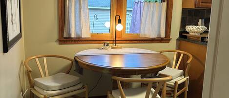 A dining area with a round wooden table and four chairs, a window with curtains, and a kitchen area with wooden cabinets.