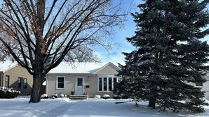 Une maison d’habitation avec une pelouse recouverte de neige, un grand arbre à feuilles persistantes et un ciel dégagé.