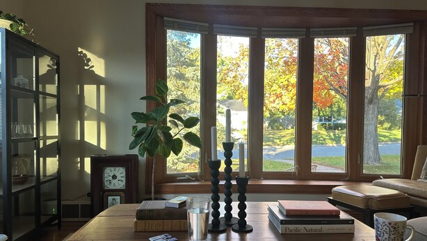 Une pièce avec une table en bois, des livres, un verre d’eau, une plante et une vue sur les arbres aux couleurs automnales, visible à travers de grandes fenêtres.