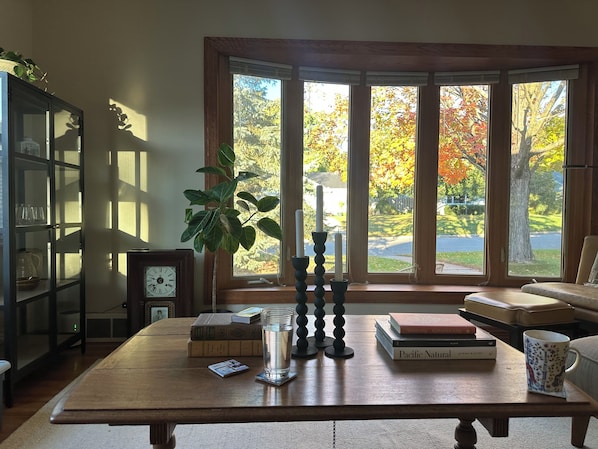 A room with a wooden table, books, a glass of water, a plant, and a view of autumn trees through large windows.