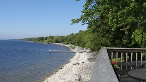 A wooden deck with a table and chairs overlooking a pebble beach and calm water.