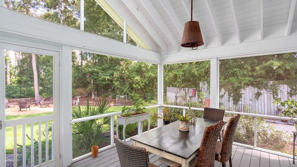 A covered patio with a table and chairs, surrounded by large windows offering views of a garden and backyard.