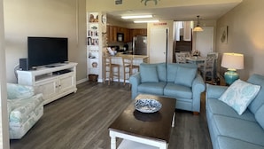 Una sala de estar con televisor, sofá, mesa de centro y un área de cocina al fondo.