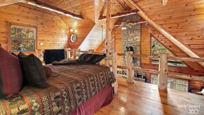 A cozy wooden cabin bedroom with a loft bed, a stone fireplace, and a balcony with a wooden railing.