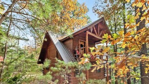 A wooden cabin with a steeply pitched roof, surrounded by trees and a stone foundation.