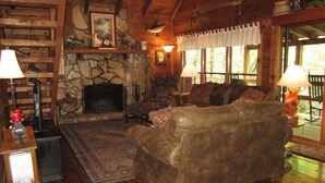 A cozy living room with a stone fireplace, wooden walls, and a large window. - Waterfront Cabin on Trout Stream W/ Hot Tub, & Internet (BLUE RIDGE)