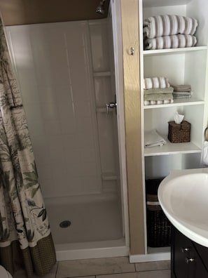 Un baño con ducha envuelta en vidrio, un lavabo blanco y estantes con toallas y artículos de aseo.