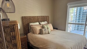 A bedroom with a woven headboard, a bed with multiple pillows, a bedside table, and a window with blinds.