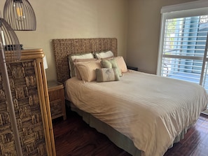 Un dormitorio con cabecera de mimbre, una cama con muchas almohadas, una mesita de noche y una ventana con persianas.