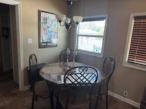 Un coin repas comprenant une table ronde, quatre chaises, un luminaire fixé au mur, une fenêtre avec des stores et une affiche encadrée accrochée au mur.