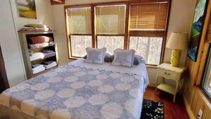 Une chambre avec un grand lit, des poutres en bois, une fenêtre avec des stores, une table de chevet avec une lampe et une étagère avec des couvertures pliées.