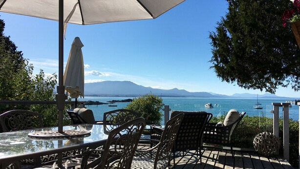 Une terrasse en bois avec des sièges d’extérieur, comprenant des chaises et une table, donnant sur une étendue d’eau où l’on aperçoit des bateaux et, au loin, des montagnes.