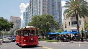 A red trolley on a city street with tall buildings, palm trees, and outdoor seating areas.