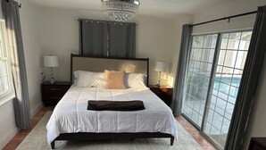 A bedroom with a large bed, two bedside tables with lamps, a chandelier, a view of a swimming pool through a sliding door, and a window with curtains.