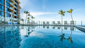 Ein klarer, blauer Infinity-Pool mit Liegestühlen und Palmen, der ein modernes Gebäude mit Balkonen widerspiegelt.