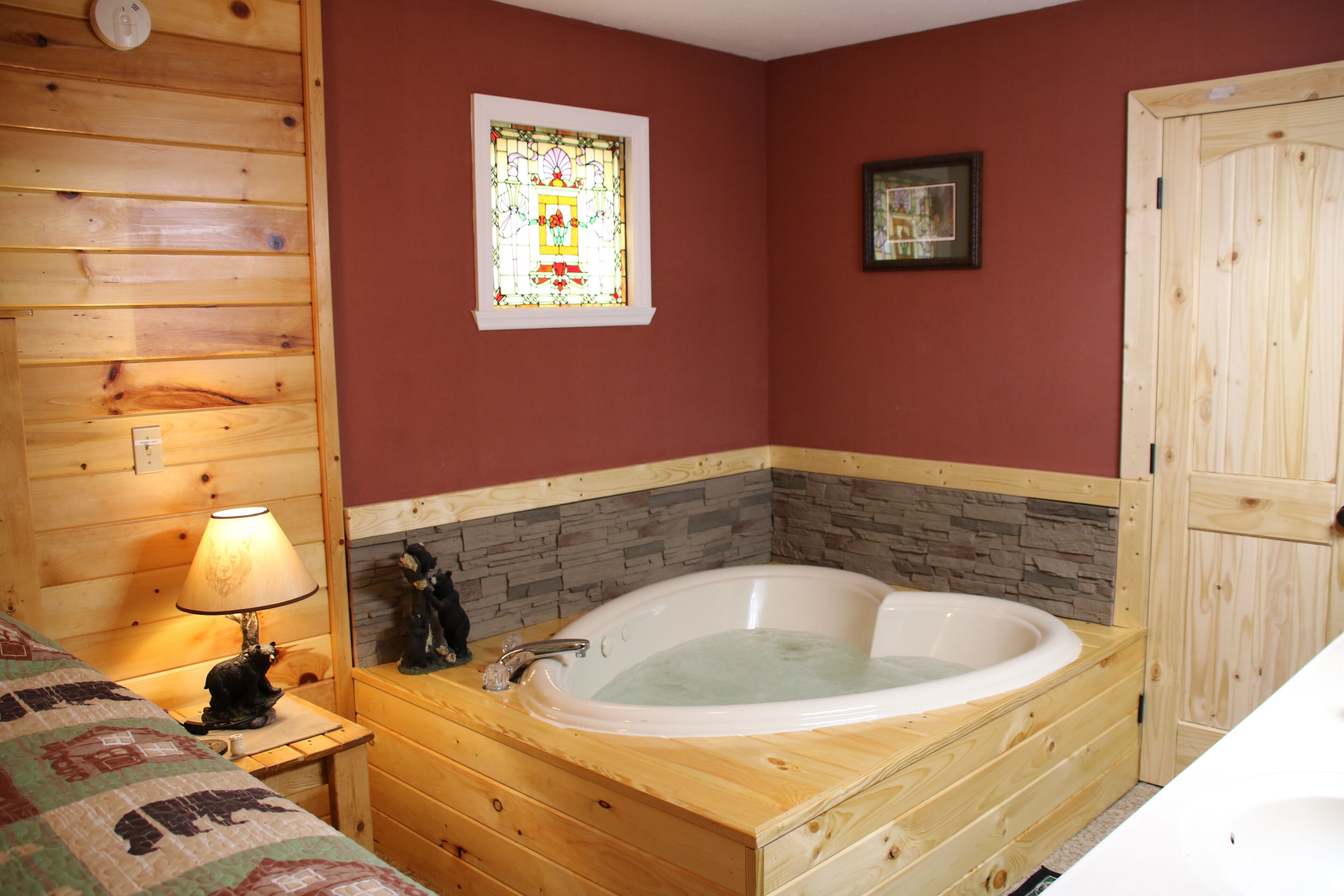 Interior de una cabaña de madera con una bañera grande de madera, una mesita de noche con lámpara, una cama acolchada y un ventanal de vitral.