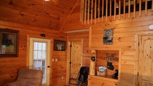 Ein Blockhaus-Interieur mit Holzwänden, Holzbalkendecke, Schaukelstuhl, gepolstertem Sessel, kleinem Tisch und Fenster mit Jalousien.