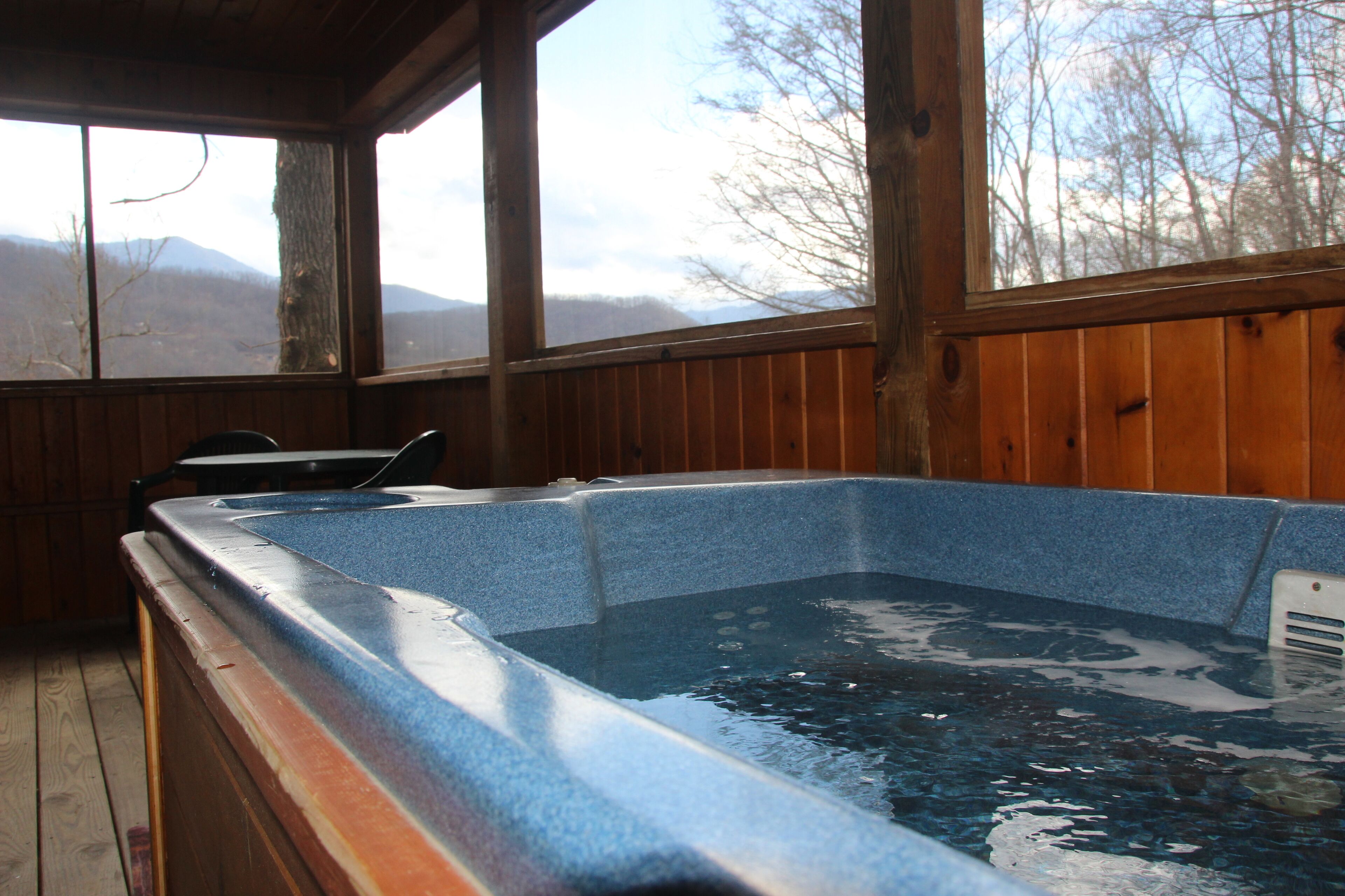 A hot tub with a wooden deck and a view of mountains through large windows.