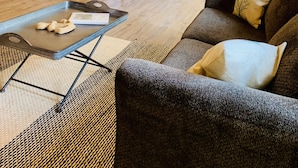 Een gezellige woonkamer met een bank en een salontafel, een houten plafond en een keuken met witte kasten en een raam.