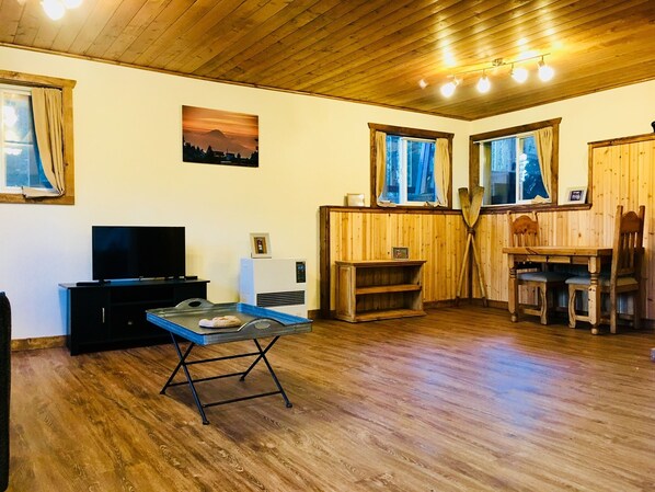 A cozy cabin interior with a wooden ceiling, a television, a small table, and a dining area with chairs. - Seldovia House - Lost Lake (Seward)