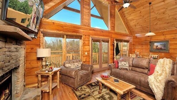 A cozy cabin interior with a stone fireplace, wooden floors, a large couch, and a ceiling fan. - Misty Mornings: Rustic Resort Cabin - Pet Friendly - New Hot Tub (Sevierville)