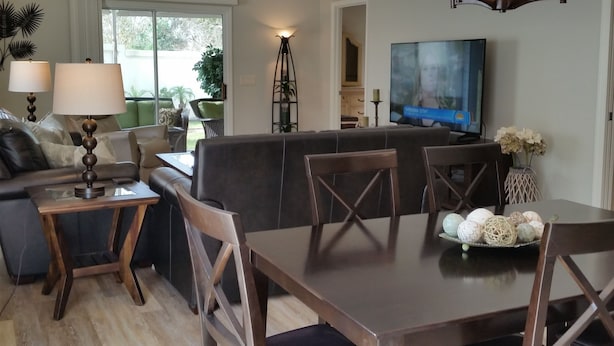 A modern living room with a dark brown sofa, a wooden coffee table, and a dining table with chairs. There is a TV mounted on the wall, a ceiling fan, and a floor lamp. - Walk to Lake Sumter Landing! Golf cart included! (The Villages)