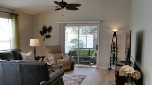 A living room with a sofa, armchair, and a glass coffee table. There is a ceiling fan, a lamp, and a vase with flowers. - Walk to Lake Sumter Landing! Golf cart included! (The Villages)