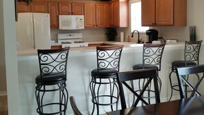 A kitchen with wooden cabinets, a white refrigerator, a microwave, a stove, and a sink. There are four bar stools with metal frames and black seats. - Walk to Lake Sumter Landing! Golf cart included! (The Villages)