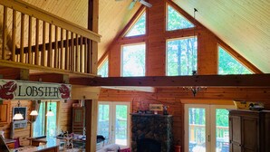 A spacious cabin with a high ceiling, wooden beams, a stone fireplace, and large windows offering views of the surrounding forest.