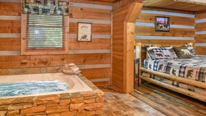Ein Interieur einer Blockhütte mit einer Stein-Whirlpool-Badewanne, einem Bett mit Tier-Dessin und einem gerahmten Bild an der Wand.