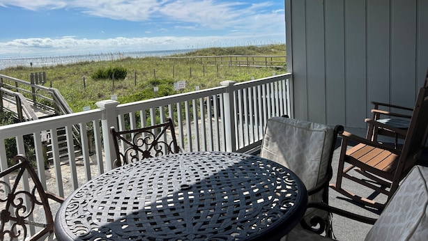 A balcony with a round metal table and two chairs, overlooking a grassy area with a fence and a view of the ocean. - Relax Oceanfront at our 2-bedroom Oceanfront condo in Carolina Beach (Carolina Beach)