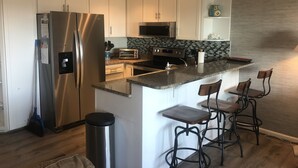 A modern kitchen with white cabinets, a stainless steel refrigerator, a microwave, and a built-in oven. There are two bar stools and a trash can. - Relax Oceanfront at our 2-bedroom Oceanfront condo in Carolina Beach (Carolina Beach)