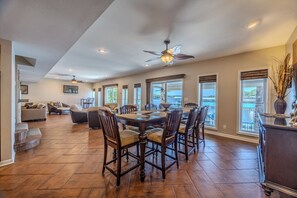 A spacious dining area with a large wooden table and chairs, a ceiling fan, and large windows offering views of the outdoors.