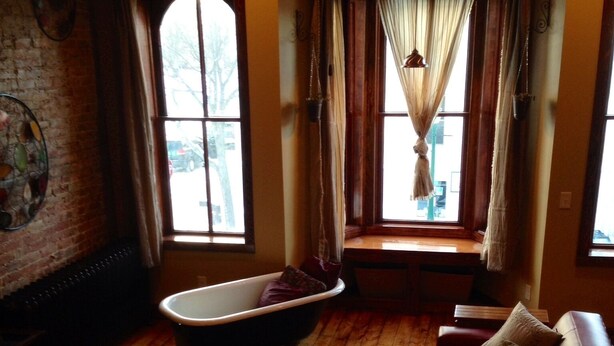A room with a red leather sofa, a vintage bathtub, and large windows with curtains. - The Nest: Beautiful Apartment Style Lodging In The Heart Of Downtown Decorah. (Decorah)