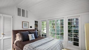 Ein Schlafzimmer mit einem großen Bett, weißen Fensterläden, einem gerahmten Bild und Blick auf Grün hinter großen Fenstern.