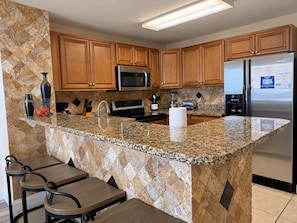 A kitchen with granite countertops, wooden cabinets, stainless steel appliances, and a bar area with stools.