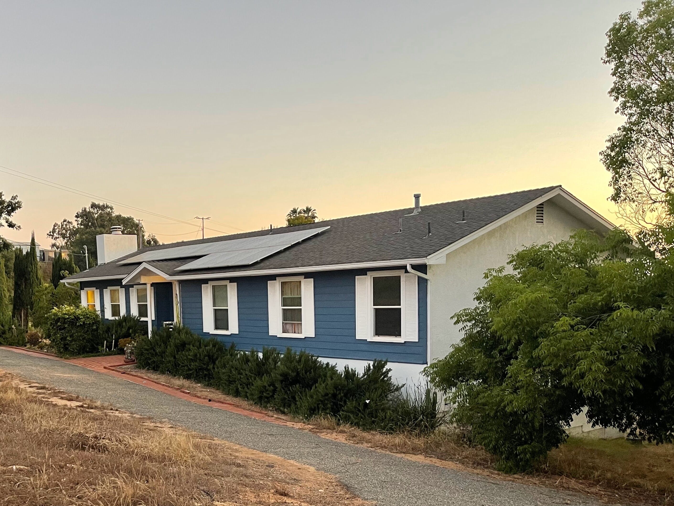 A single-story house with a grey roof and blue siding, solar panels on the roof, and a driveway leading to the front door.