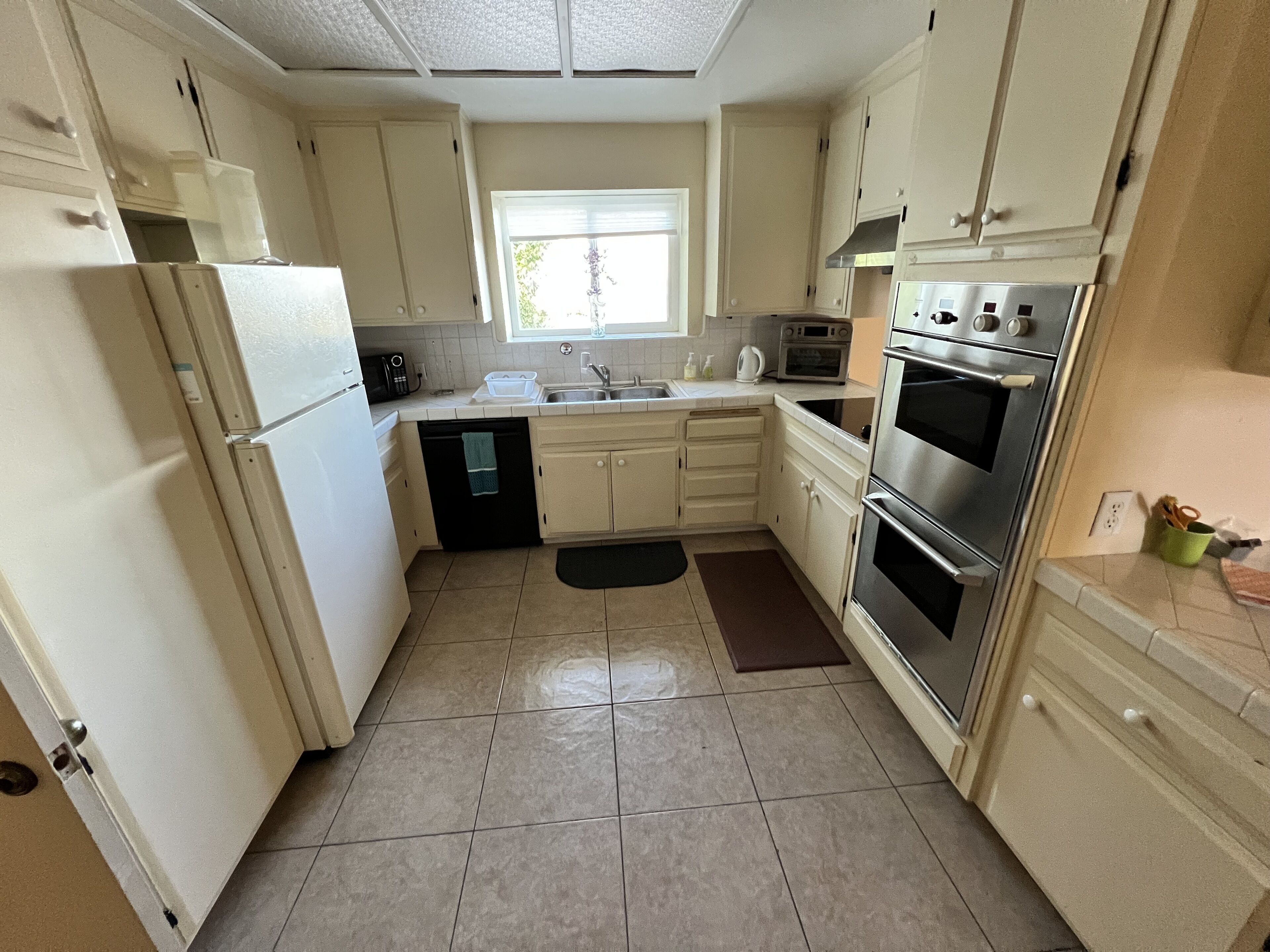 A compact kitchen with white cabinets, a stainless steel refrigerator, a built-in oven, and a window above the sink.