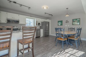 A modern kitchen with white cabinets, a stainless steel refrigerator, and a dining area with wooden chairs. - The Kure for all your troubles. (Kure Beach)